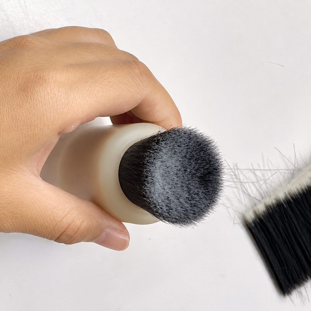 A hand holds a small makeup brush with dense black bristles and a cream-colored handle against a plain white background, with another brush partially visible on the right.